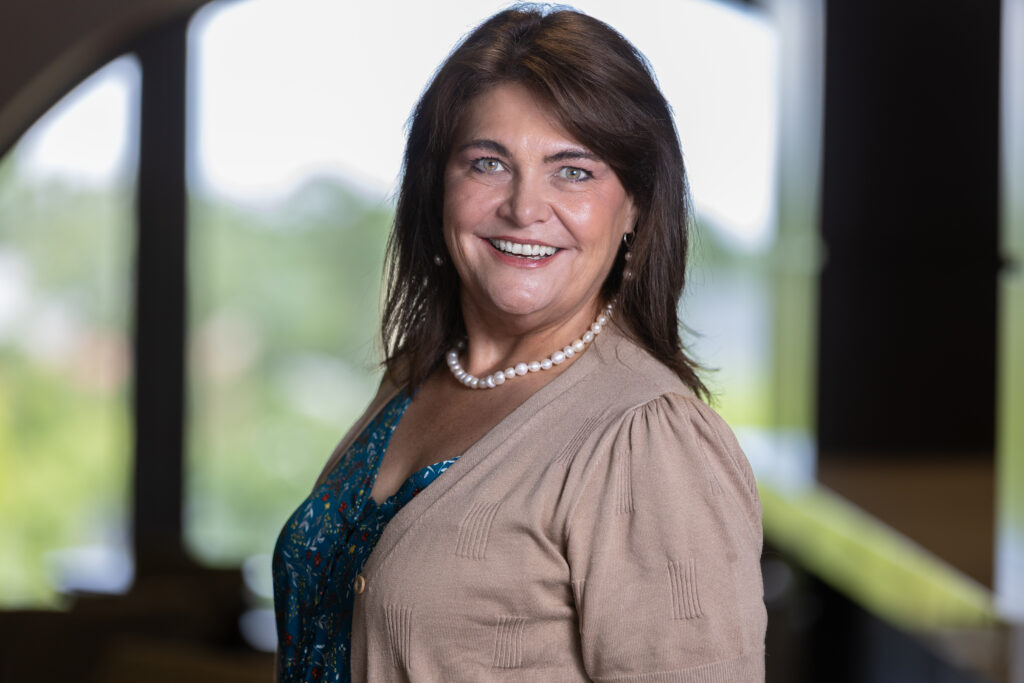 A smiling woman with long brown hair wearing a tan cardigan, teal patterned dress, and pearl necklace stands indoors with a blurred, sunlit background.
