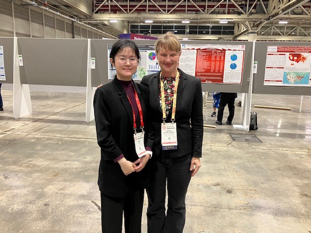 Two women stand smiling in front of scientific posters at a conference. Both wear business attire and conference badges. The background shows other posters and attendees in a large exhibition hall.