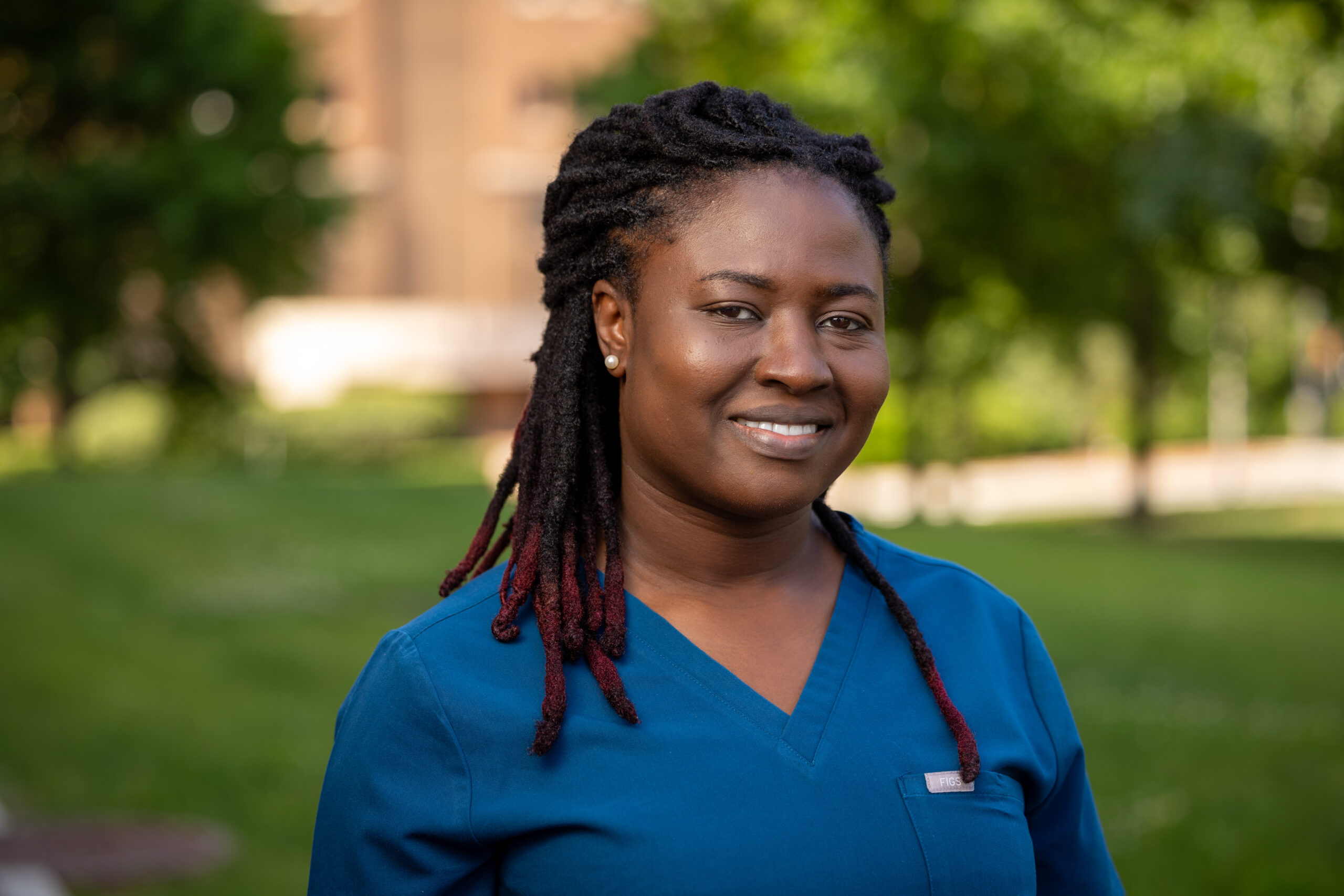 A woman with long braided hair wearing blue medical scrubs smiles while standing outdoors in a green, sunlit park.