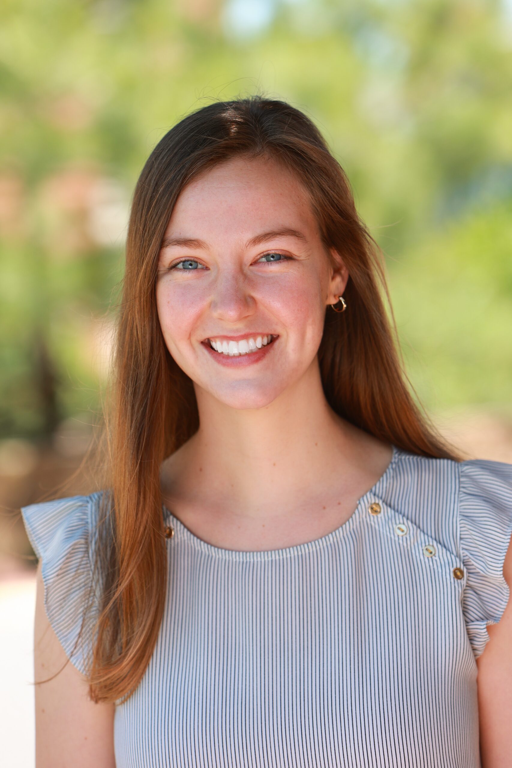 A young woman with long brown hair, wearing a light-colored, short-sleeve top with button details, smiles outdoors with a blurred green background.