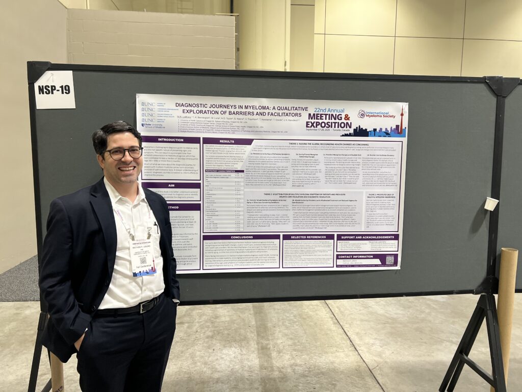 A man in a suit and glasses stands smiling next to a scientific poster on a display board at a conference. The poster is titled Diagnostic Journeys in Myeloma and the event is labeled as the 22nd Annual Meeting & Exposition.