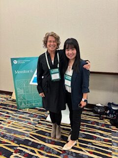 Two women stand smiling together at a conference. Both wear name badges and business attire. Behind them is a sign that reads Monitor 6 and a patterned carpet covers the floor.