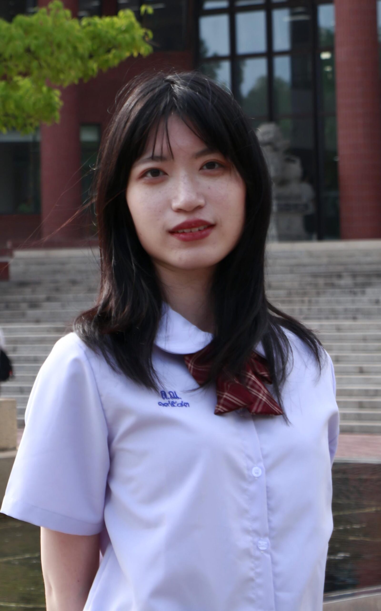 A young woman with long dark hair stands outdoors in front of stone steps and a brick building, wearing a white blouse and a red plaid bow tie. She is looking at the camera with a slight smile.