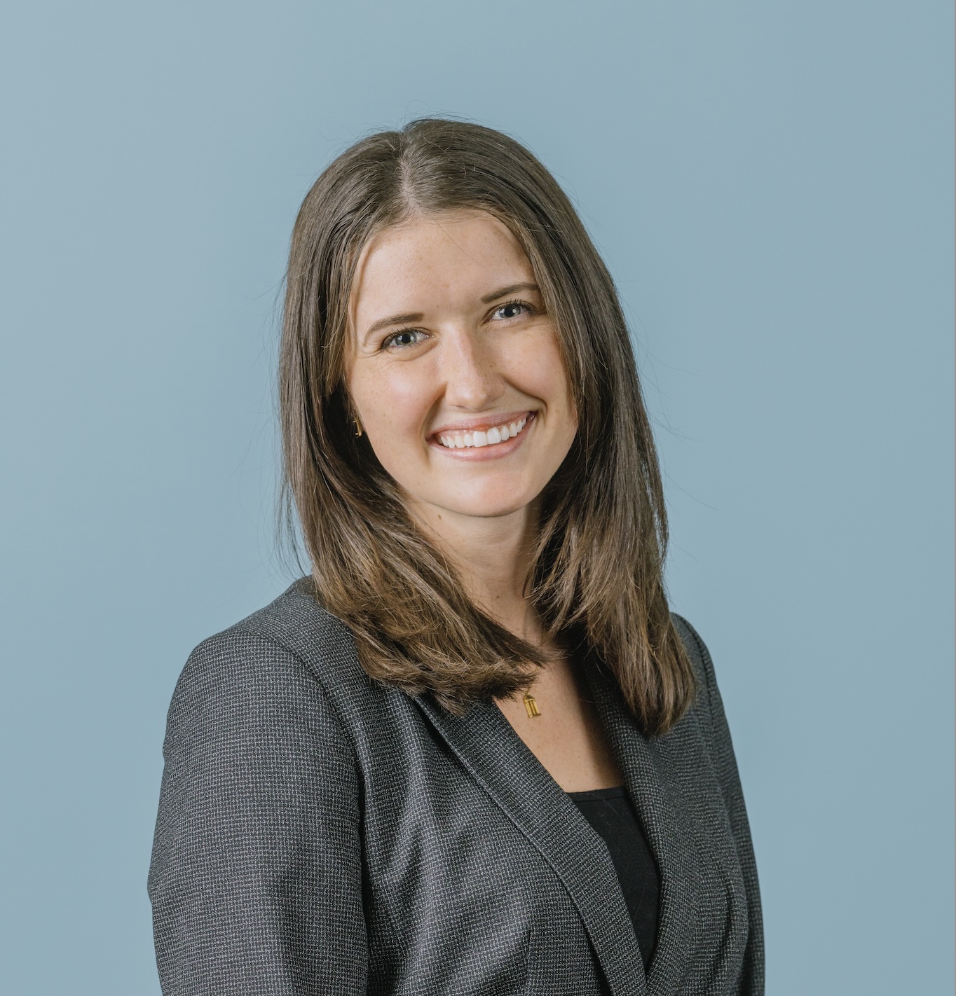 A woman with straight brown hair wearing a dark blazer and black top smiles at the camera in front of a plain light blue background.