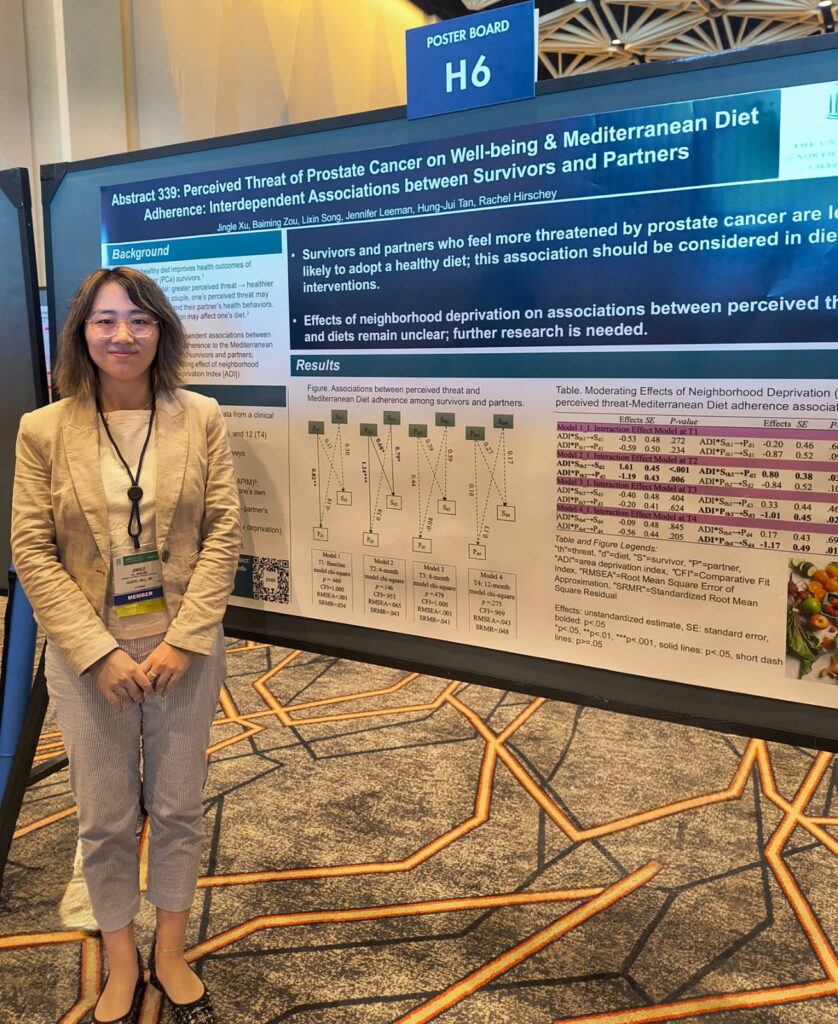 A woman stands beside a scientific poster about prostate cancer, well-being, and Mediterranean diet adherence at a conference. The poster includes text, tables, and charts. The setting appears to be a conference hall.