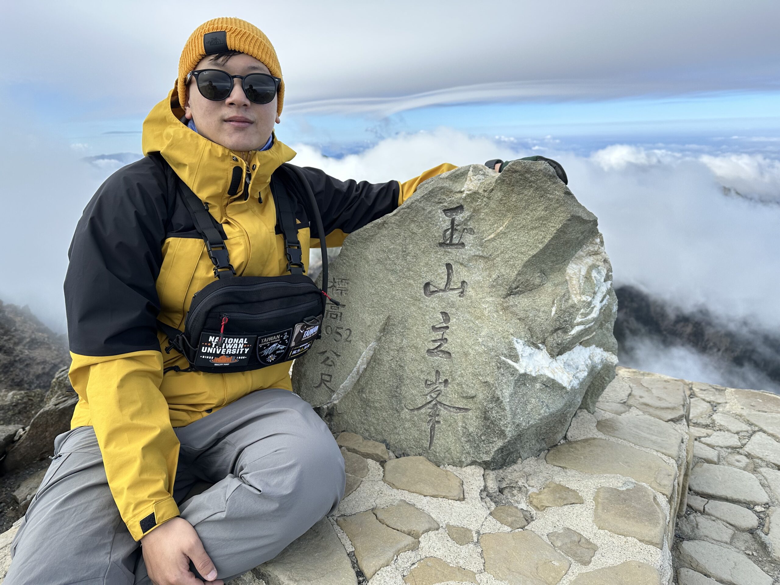 A person in a yellow and black jacket, gray pants, and sunglasses poses next to a large rock with Asian characters engraved on it at a mountain summit, surrounded by clouds and a stone platform.