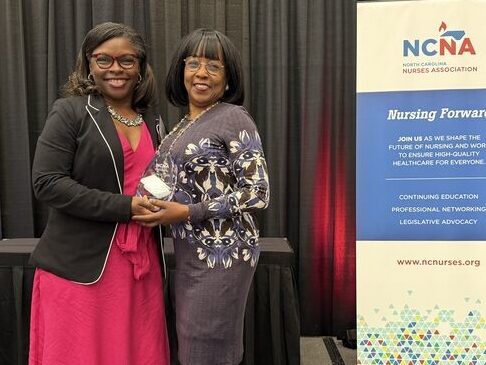 Two women stand smiling and holding an award on a stage next to an NCNA Nursing Association sign, with a black curtain backdrop and green plant to the right. One wears a pink dress and black jacket; the other wears a patterned dress.