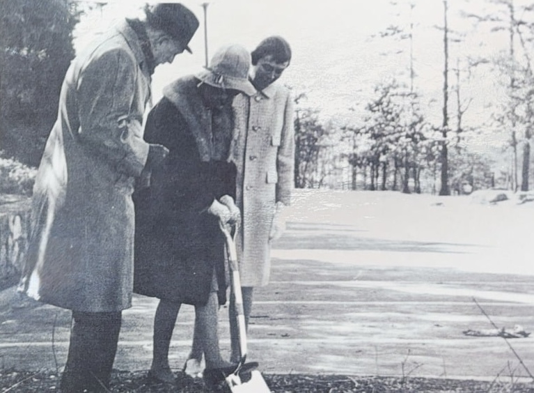 Three people in vintage clothing participate in a groundbreaking ceremony outdoors; one person in a hat and fur-collared coat uses a shovel, while the other two stand nearby watching. Trees and sunlight are visible in the background.