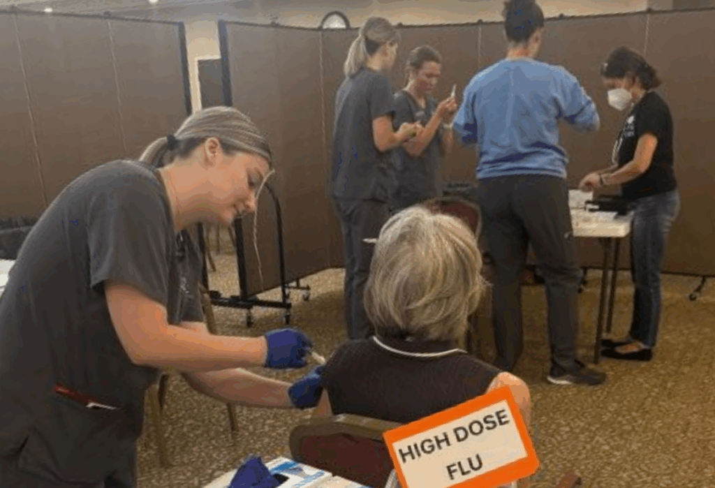 A healthcare worker administers a flu shot to an elderly person seated near a sign that reads HIGH DOSE FLU, while other medical staff prepare supplies in the background.