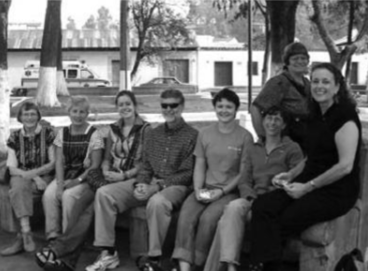 A group of eight adults, mostly women, sit closely together on an outdoor bench, smiling at the camera. Trees and buildings are visible in the background. The image is in black and white.