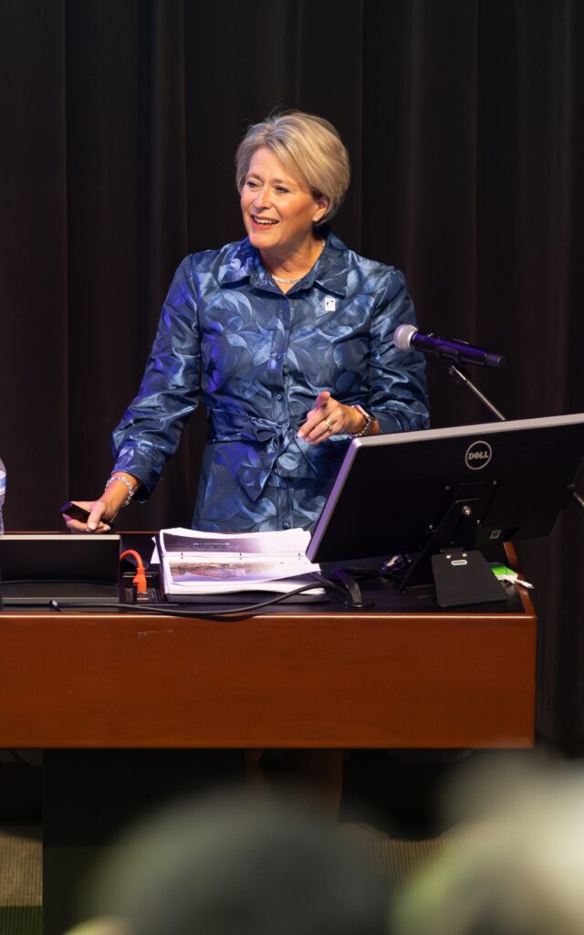 Dean Val Howard in blue patterned jacket stands at a lectern, smiling while delivering the 2025 State of the School address for the UNC School of Nursing. She is holding a pointer and standing behind a computer monitor and microphone, with papers and a water bottle on the desk. Dark curtains are in the background.