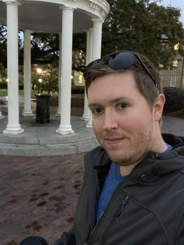 A man with short brown hair and sunglasses on his head takes a selfie outdoors in front of a white columned gazebo on a brick walkway, with trees and buildings visible in the background.