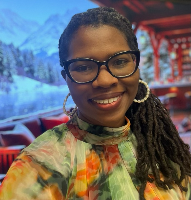 Smiling woman with glasses and long braided hair wears a floral top. She stands indoors in front of a backdrop featuring snowy mountains, pine trees, and a cozy lodge with red furniture.