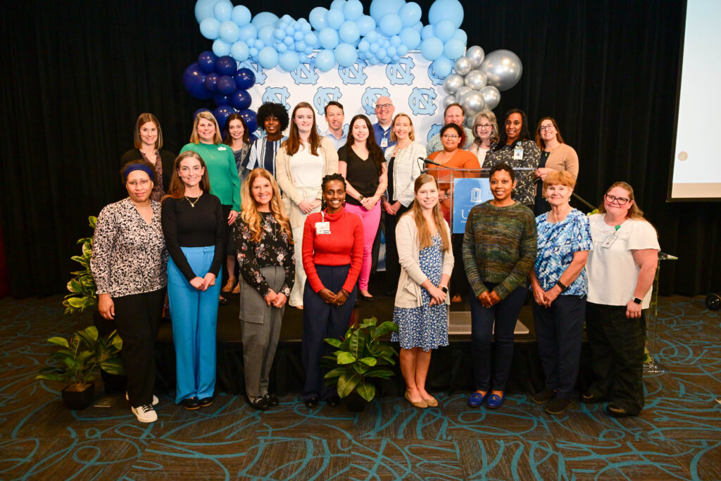 A diverse group of people pose together on a stage decorated with blue and white balloons and potted plants, all smiling at the camera in a celebratory indoor setting.