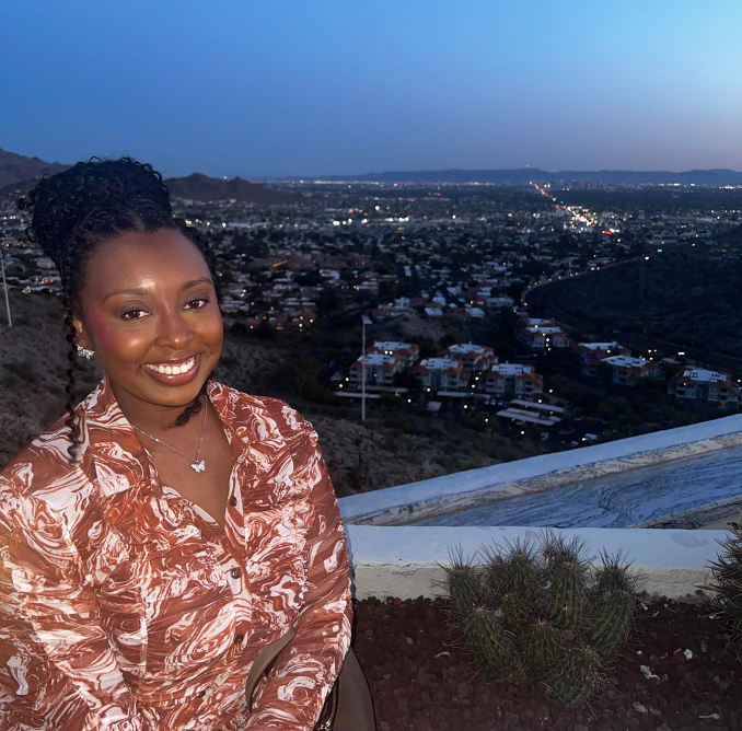 A woman in a patterned brown and white blouse smiles at the camera, sitting outdoors at dusk with a cityscape and distant mountains in the background. Small cacti and city lights are visible behind her.