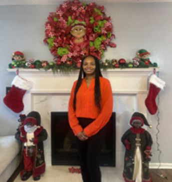 A woman in an orange top stands smiling in front of a decorated fireplace with stockings, festive figurines, and a large holiday wreath above the mantel.