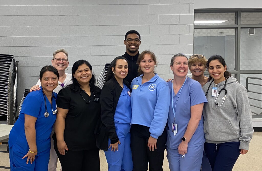 A group of ten smiling healthcare workers, some in scrubs and others in casual clothing, stand together indoors in front of a gray wall and stacked chairs.
