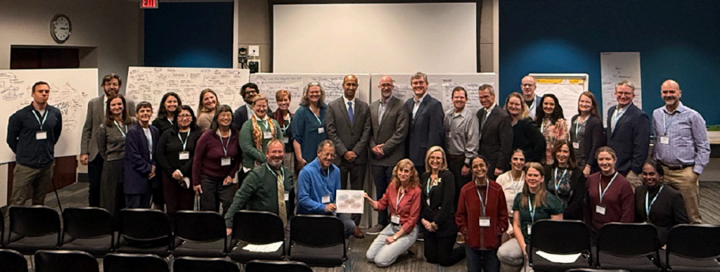 A large group of people, both seated and standing, pose for a photo in a conference room with whiteboards covered in notes and diagrams behind them. Most are wearing name badges and smiling at the camera.
