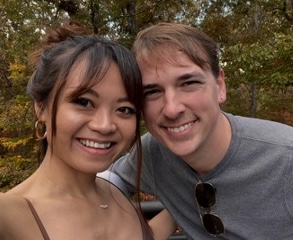 A smiling woman and man pose closely together outdoors with trees bearing autumn leaves in the background. The woman has long dark hair and wears earrings; the man wears a grey shirt and sunglasses hanging from his collar.