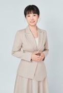 A woman with short black hair, wearing a beige suit jacket and skirt, stands against a light gray background, smiling gently with her hands clasped in front of her.