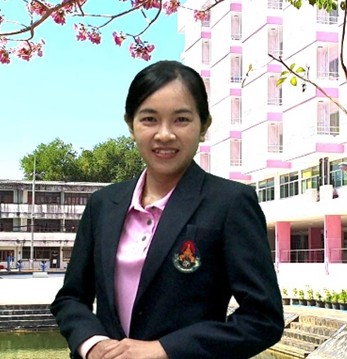 A woman in a black blazer and pink shirt stands smiling in front of a modern building with pink and white accents, with trees and blooming branches in the background.