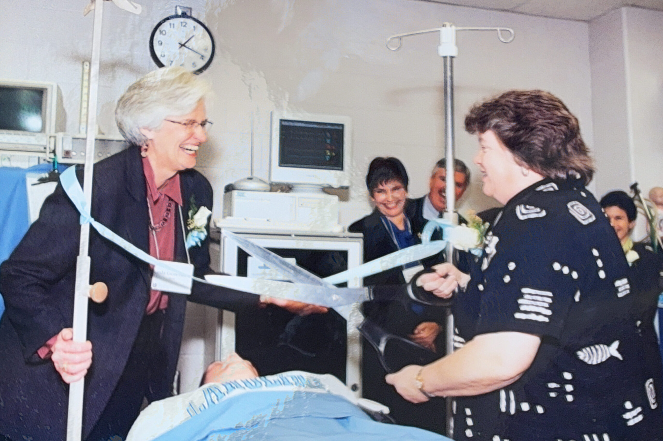 Two women smile while cutting a ribbon across a hospital bed in a medical setting, surrounded by several people, including medical staff. A clock and medical equipment are visible in the background.