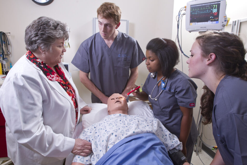 A medical instructor and three students in scrubs practice a procedure on a medical mannequin in a hospital room, with monitoring equipment visible in the background.