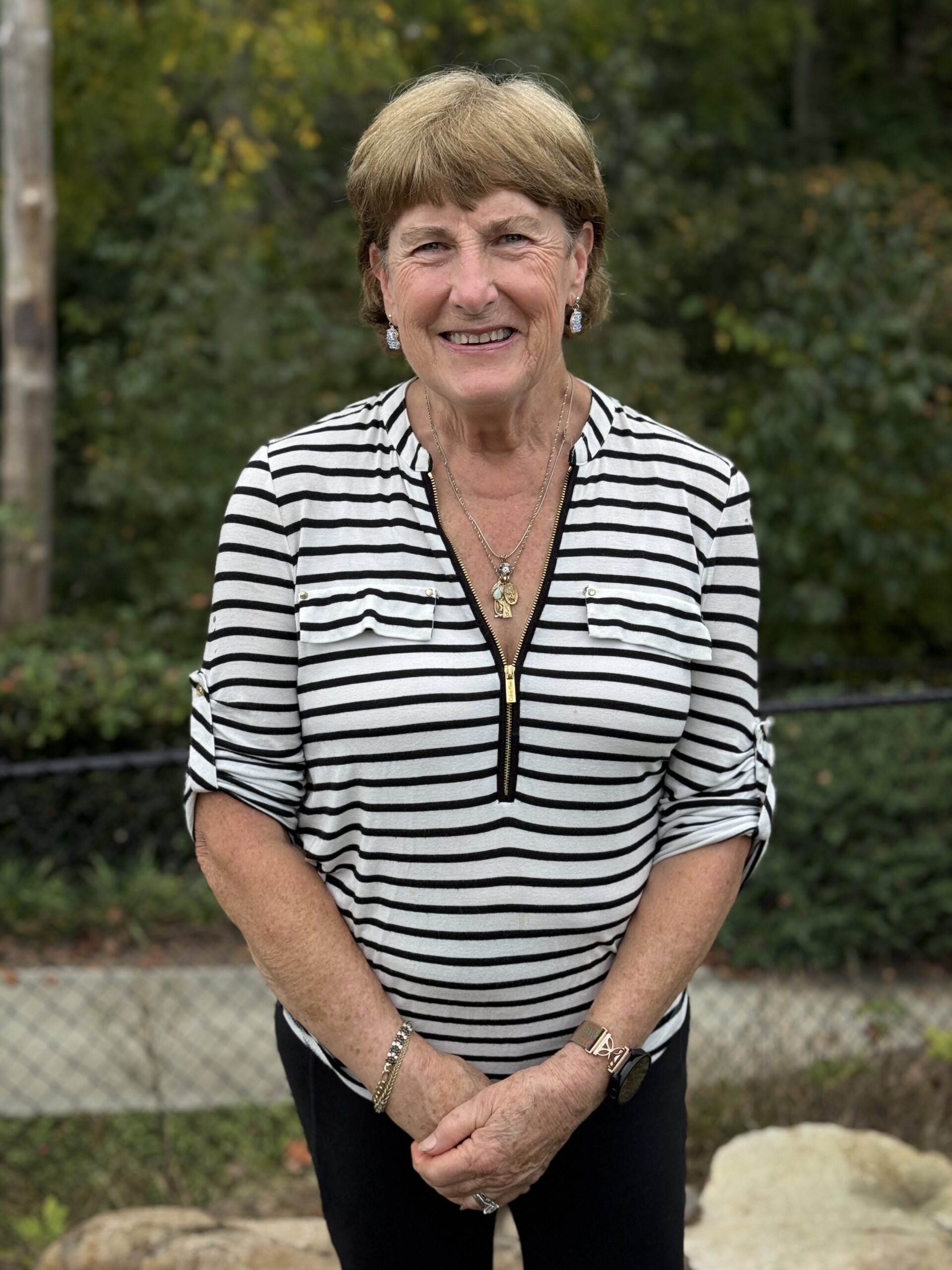 A smiling older woman with short light brown hair wearing a black and white striped shirt stands outdoors in front of greenery, rocks, and a black chain-link fence.