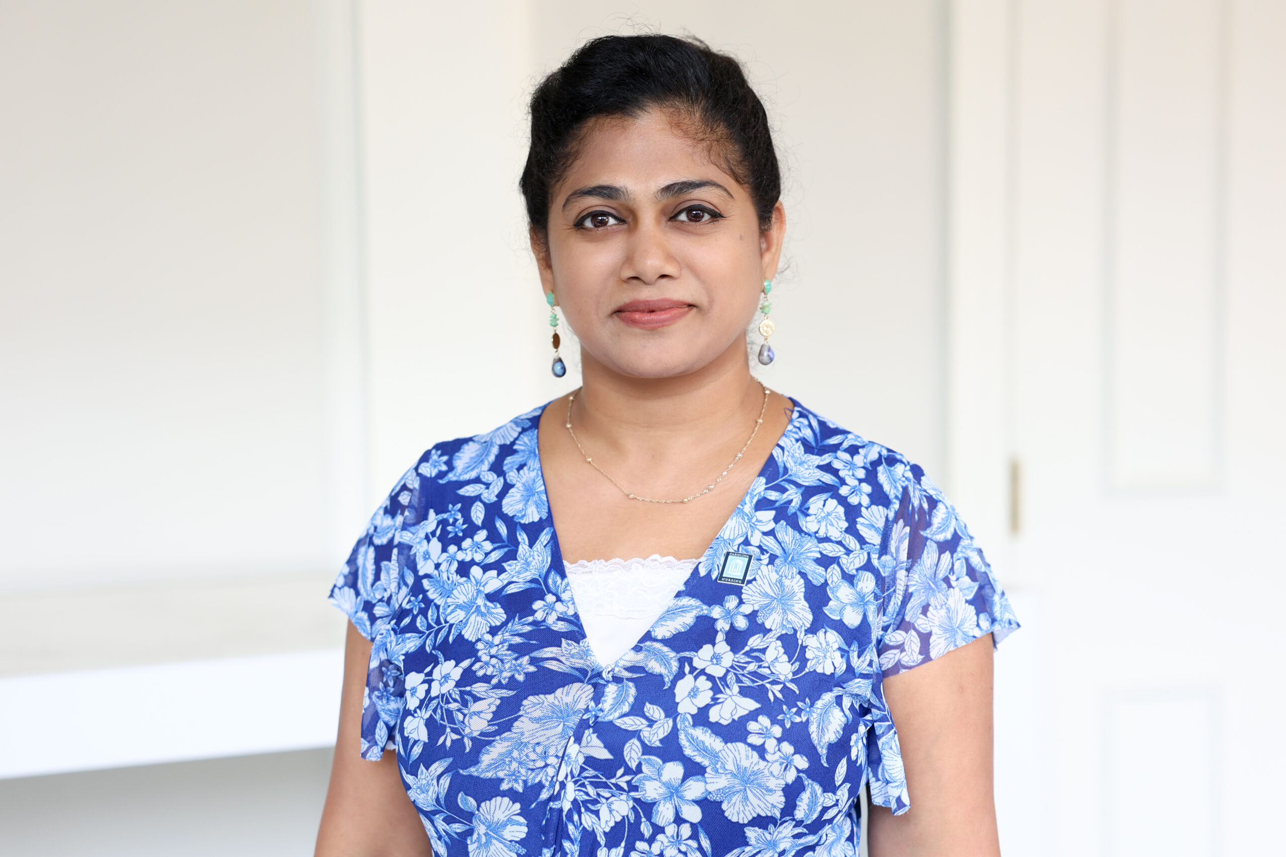 A woman with dark hair pulled back, wearing a blue floral blouse and drop earrings, stands in a bright room with white walls and a door in the background. She is looking at the camera with a neutral expression.