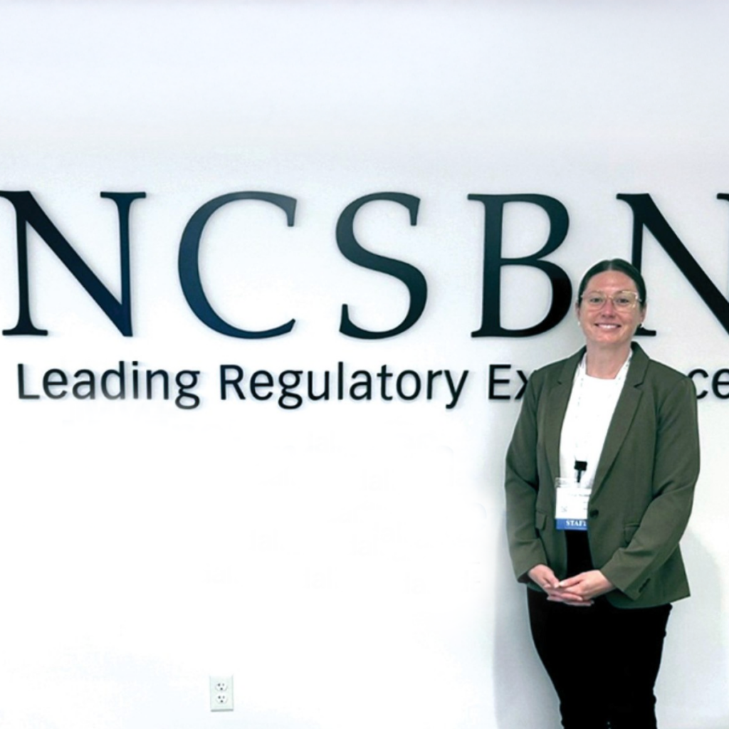 A person wearing glasses and a blazer stands smiling in front of a white wall with large letters that read NCSBN and the phrase Leading Regulatory Excellence partially visible.
