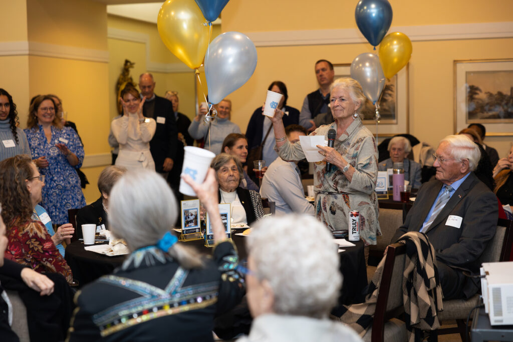 Jo-Anne Trowbridge Martin holds a microphone and a cup in a toast during the groundbreaking ceremony of the new Nursing Education Building. Others seated around tables raise cups in a toast. Blue and gold balloons decorate the room.