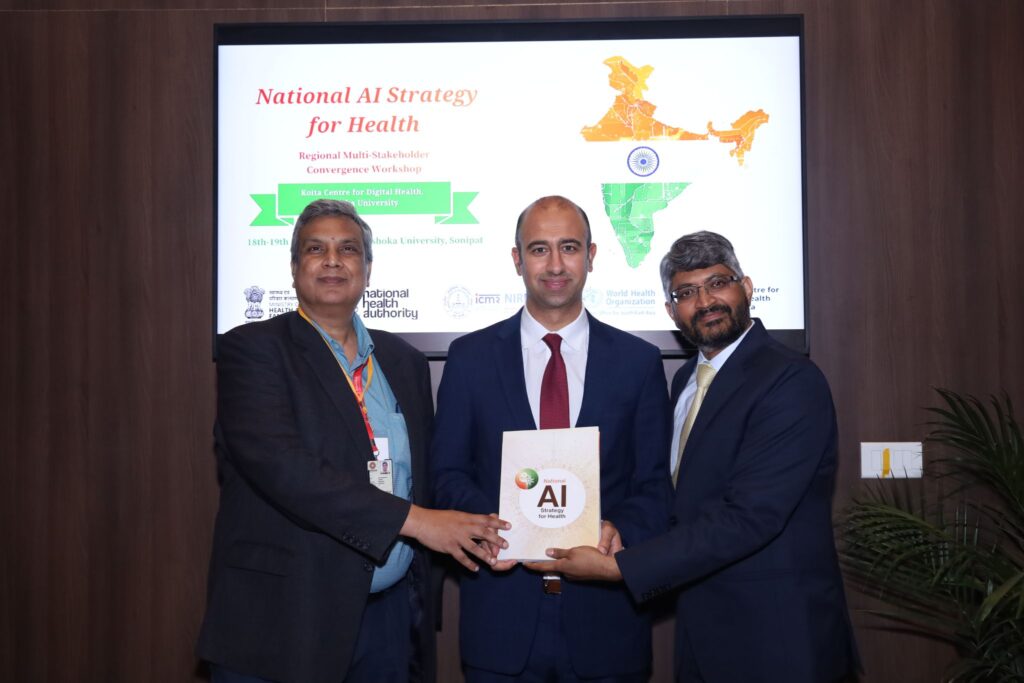Three men in formal attire stand in front of a screen displaying National AI Strategy for Health; one man in the center holds a booklet with AI on the cover as they pose for a photo at a conference or workshop event.