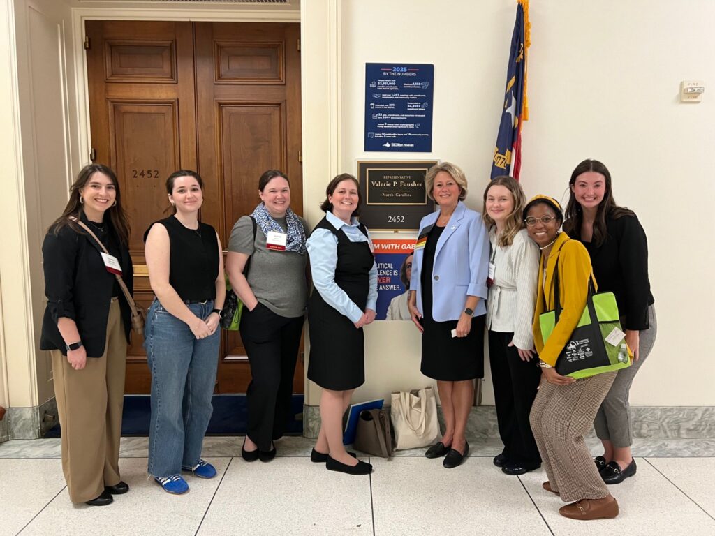 Eight women stand smiling in a hallway in front of an office door with a sign reading “Walter B. Jones Jr. 2452.” One woman holds a green tote bag; an American flag is visible behind them.