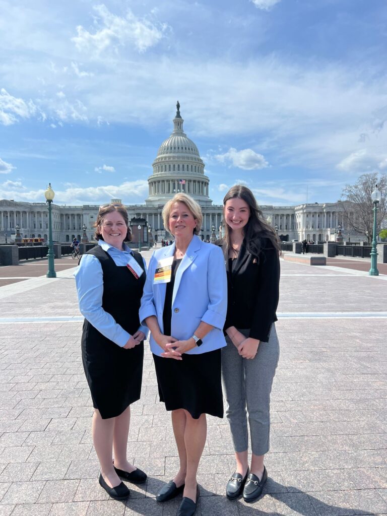 Three women stand smiling in front of the U.S. Capitol building on a sunny day. Two wear business attire with badges, and one wears a light blue blazer. The sky is mostly clear with a few clouds.
