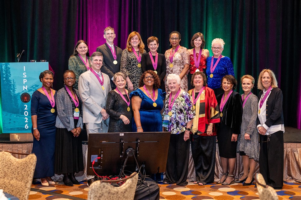 A group of sixteen people, wearing medals and formal attire, stand together and smile for a photo on stage at an ISPN 2026 event, with a podium and colorful curtains in the background.