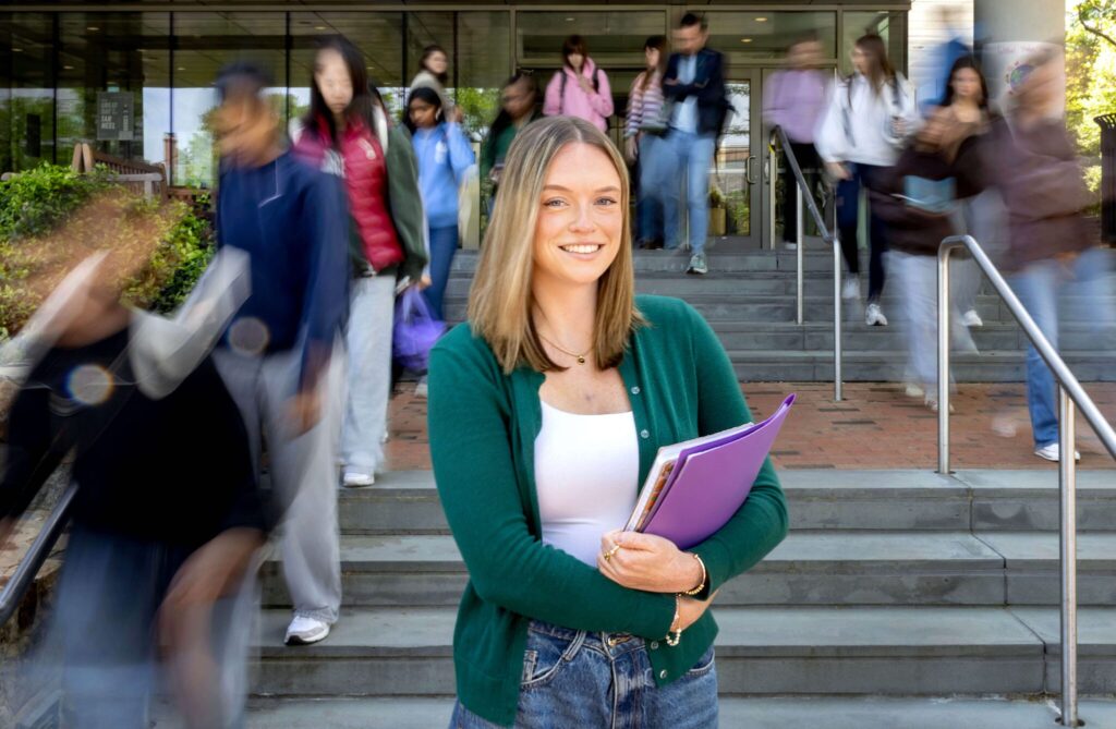 A young, white woman with blonde hair and a green sweater smiles and holds her school books in front of a staircase.