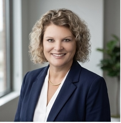 Professional portrait of Michelle Edmonds wearing a navy blazer, white blouse, and gold necklace smiling in a bright office with a window and plant in the background.