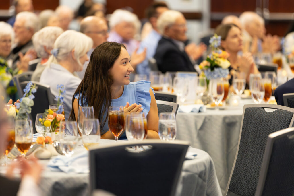 UNC School of Nursing Scholarship recipient in a light blue dress sits at a round table, smiling and clapping during the Carington Society Dinner. The room is filled with people seated at tables set with floral centerpieces and iced tea glasses.