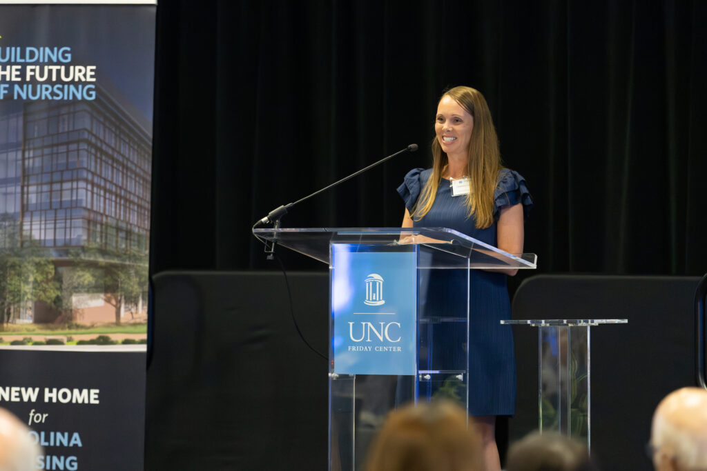 A woman stands at a clear podium with the UNC logo, smiling and speaking to an audience. Behind her is a banner that reads Building the Future of Nursing and New Home for Carolina Nursing.