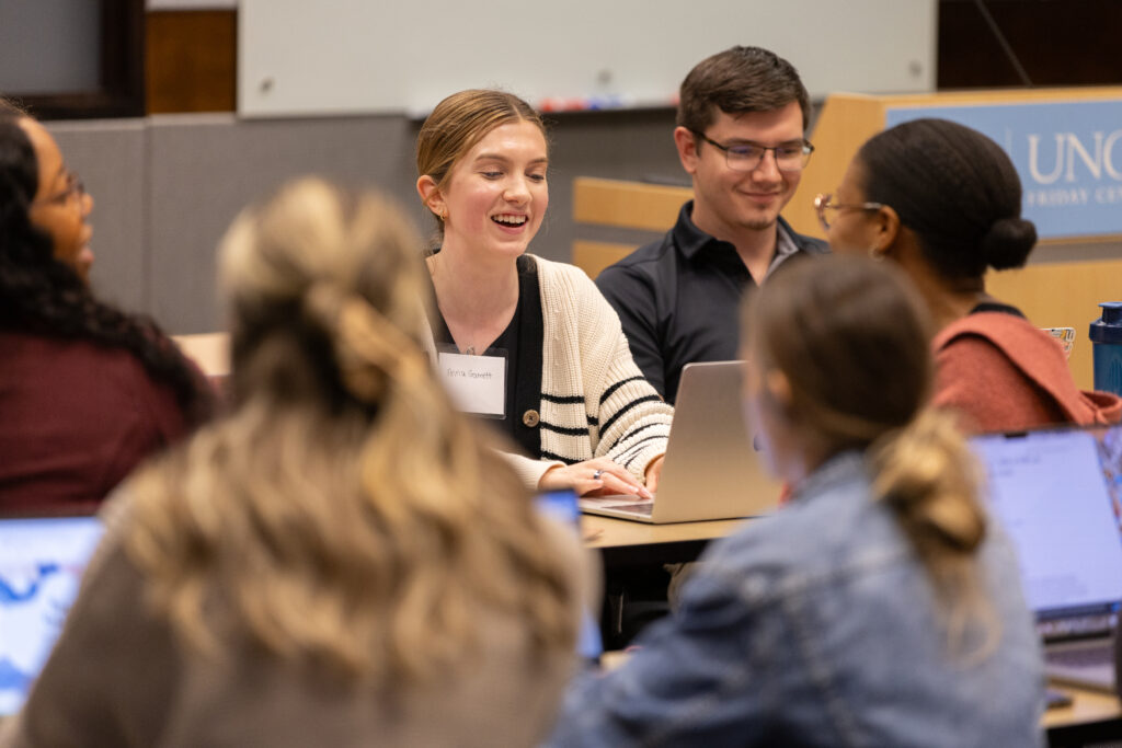 A group of UNC School of Nursing DNP students sits around a table engaged in discussion, with laptops open in front of them. One student in the center is smiling and talking. A podium with a UNC logo is visible in the background.