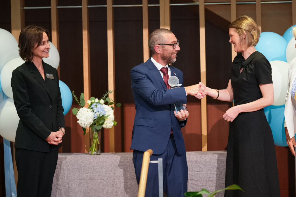 A man in a suit holding a glass award shakes hands with a woman in a black dress on stage, while another woman in a black suit stands nearby, smiling. Blue and white balloons and flowers decorate the background.