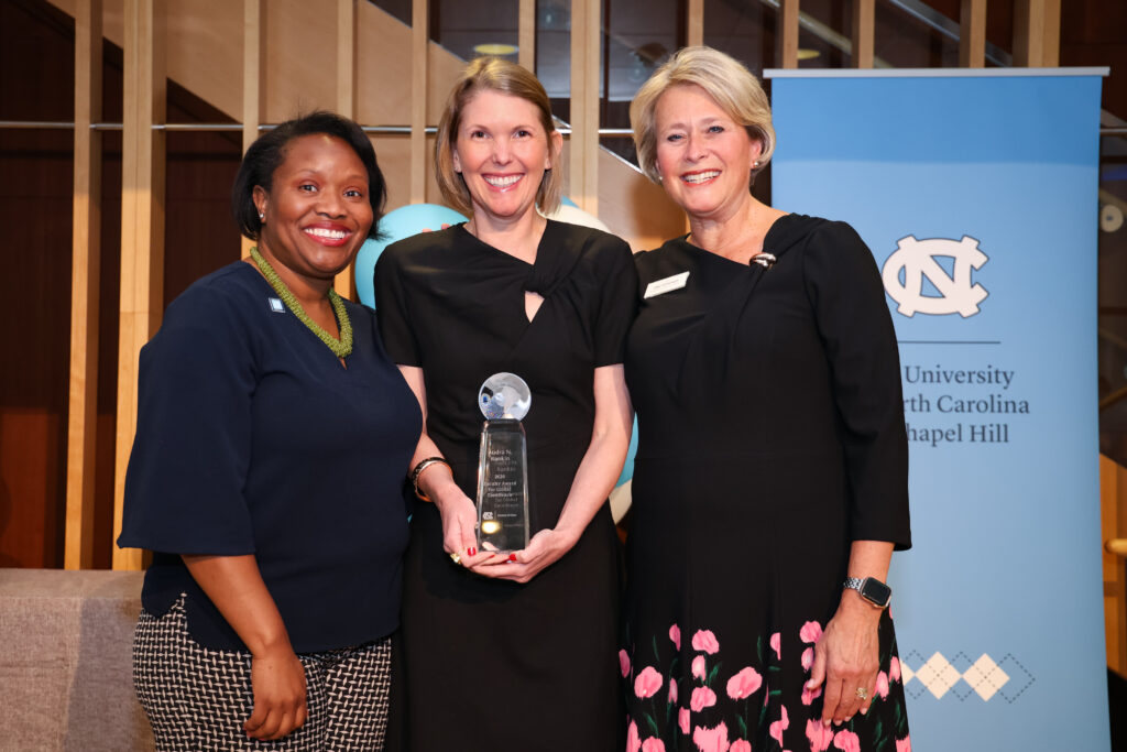 Three women stand together smiling; the woman in the center holds a glass award. They are in front of a blue University of North Carolina at Chapel Hill banner inside a well-lit building.