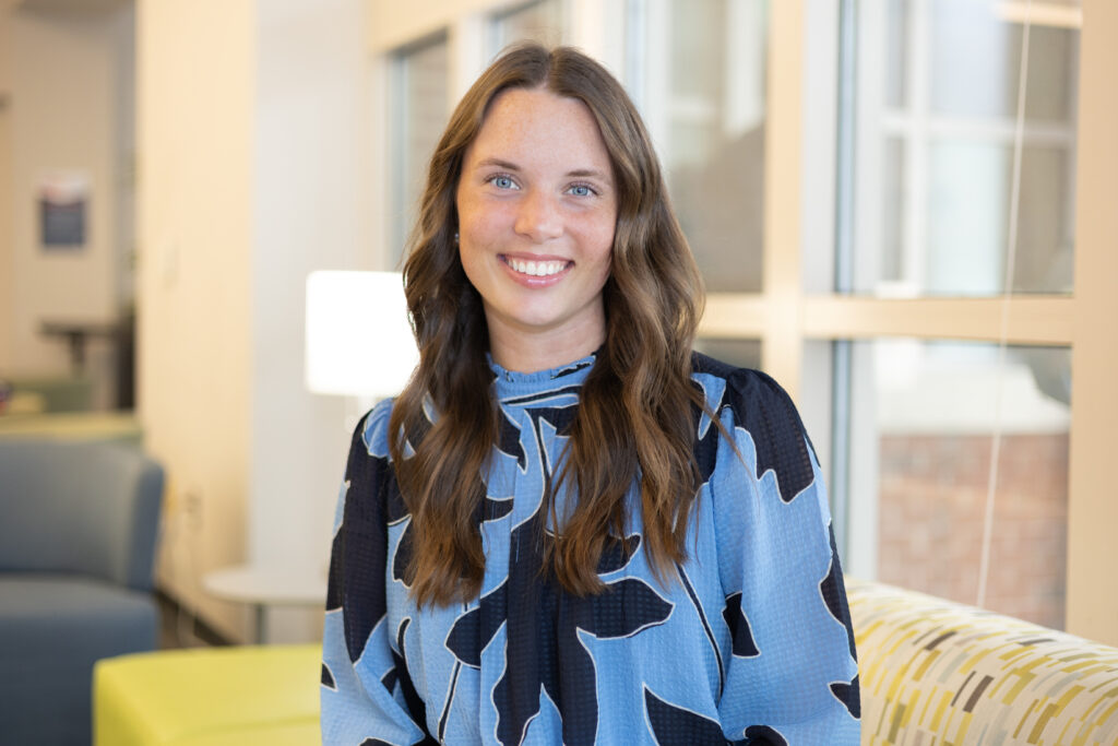 A woman with long brown hair, wearing a blue and black patterned blouse, smiles while sitting indoors on a patterned couch with large windows in the background.