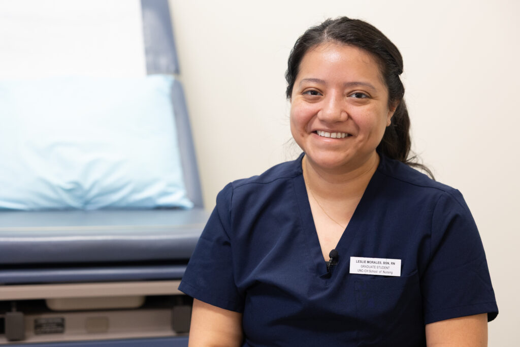 A woman in navy blue medical scrubs smiles while sitting in a clinic room next to an exam table with a blue pillow. She wears a name badge and has a microphone clipped to her shirt.