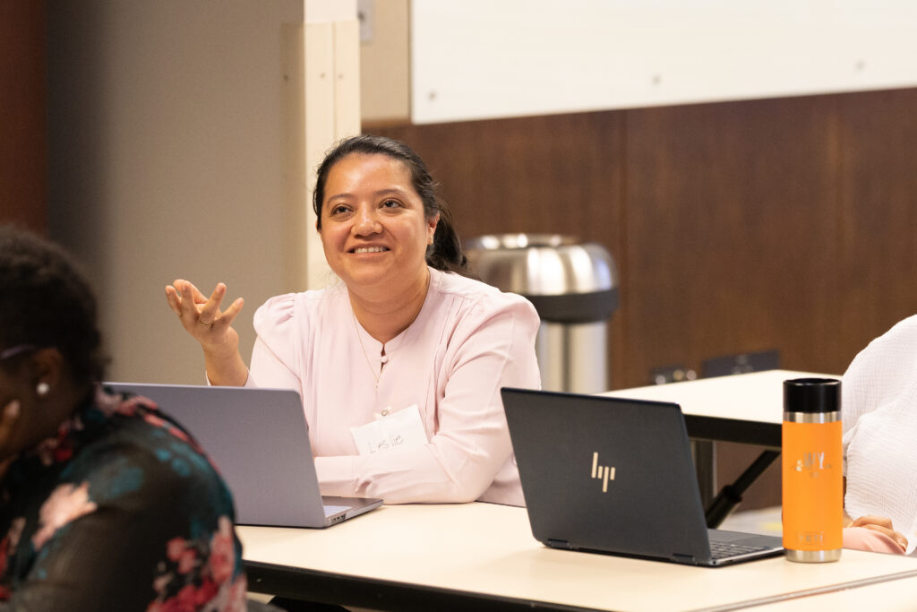 A woman wearing a light pink blouse sits at a desk with an HP laptop, smiling and gesturing with her hand during a meeting or class. Another person and an orange water bottle are also visible.
