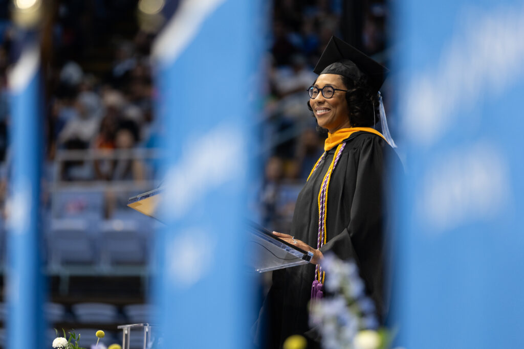 A smiling graduate in a cap and gown stands at a podium, speaking at a graduation ceremony. Blue banners frame the image, and an audience is blurred in the background.