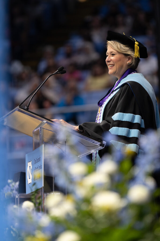 A woman in academic regalia smiles while speaking at a podium during a graduation ceremony, with blurred flowers in the foreground and an audience in the background.