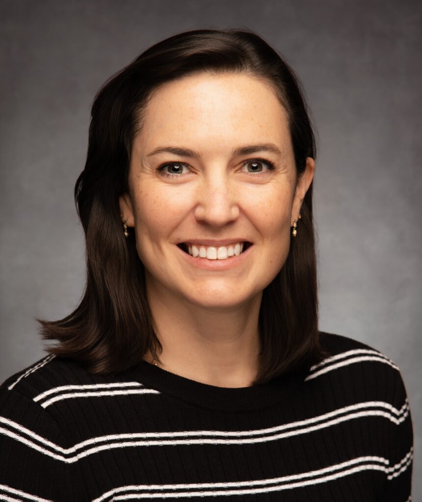 A woman with straight, shoulder-length dark brown hair smiles at the camera. She is wearing a black sweater with white horizontal stripes and gold earrings, set against a plain, dark background.