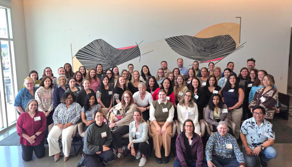 A large group of people pose together indoors in front of a wall with abstract art. Most are smiling and wearing name tags. The group includes a mix of men and women of varying ages, seated and standing in rows.