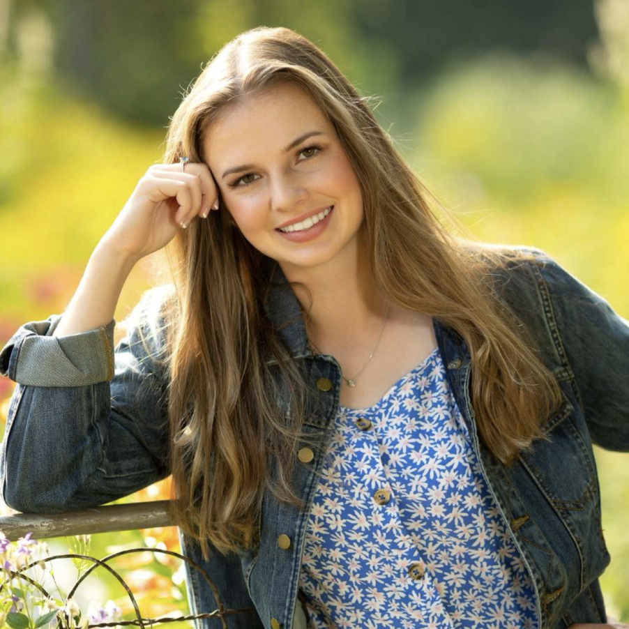 A young woman with long brown hair is smiling and leaning on a wooden fence. She is wearing a denim jacket over a blue floral top, with a blurred, sunlit garden in the background.
